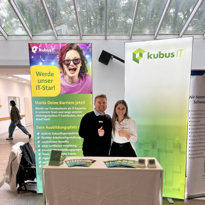 Auf dem Foto stehen ein Mann und eine Frau hinter einem Infostand von kubus IT. Beide lächeln und zeigen einen Daumen nach oben. Hinter ihnen befinden sich zwei große Roll-up-Banner mit dem kubus IT Logo und Informationen zu Ausbildungs- und Studienmöglichkeiten. Auf dem Tisch liegen mehrere Flyer aus. Der Stand befindet sich in einer hellen Halle mit Glasdach.