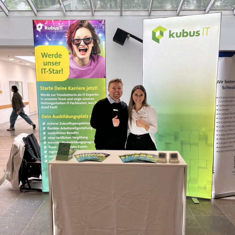 Auf dem Foto stehen ein Mann und eine Frau hinter einem Infostand von kubus IT. Beide lächeln und zeigen einen Daumen nach oben. Hinter ihnen befinden sich zwei große Roll-up-Banner mit dem kubus IT Logo und Informationen zu Ausbildungs- und Studienmöglichkeiten. Auf dem Tisch liegen mehrere Flyer aus. Der Stand befindet sich in einer hellen Halle mit Glasdach.