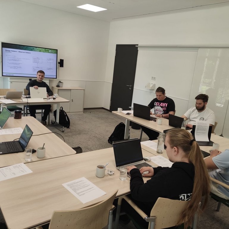 Das Bild zeigt eine Gruppe von Auszubildenden der kubus IT in einem modernen Seminarraum. Sie sitzen mit Laptops an Tischen und arbeiten konzentriert während eines Lehrgangs zur objektorientierten Programmierung. Im Hintergrund ist ein Dozent zu sehen, der Inhalte auf einem großen Bildschirm präsentiert. Die Atmosphäre wirkt fokussiert und praxisnah.