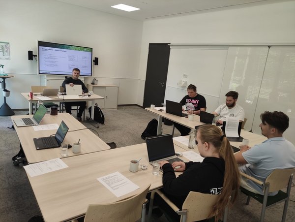 Das Bild zeigt eine Gruppe von Auszubildenden der kubus IT in einem modernen Seminarraum. Sie sitzen mit Laptops an Tischen und arbeiten konzentriert während eines Lehrgangs zur objektorientierten Programmierung. Im Hintergrund ist ein Dozent zu sehen, der Inhalte auf einem großen Bildschirm präsentiert. Die Atmosphäre wirkt fokussiert und praxisnah.
