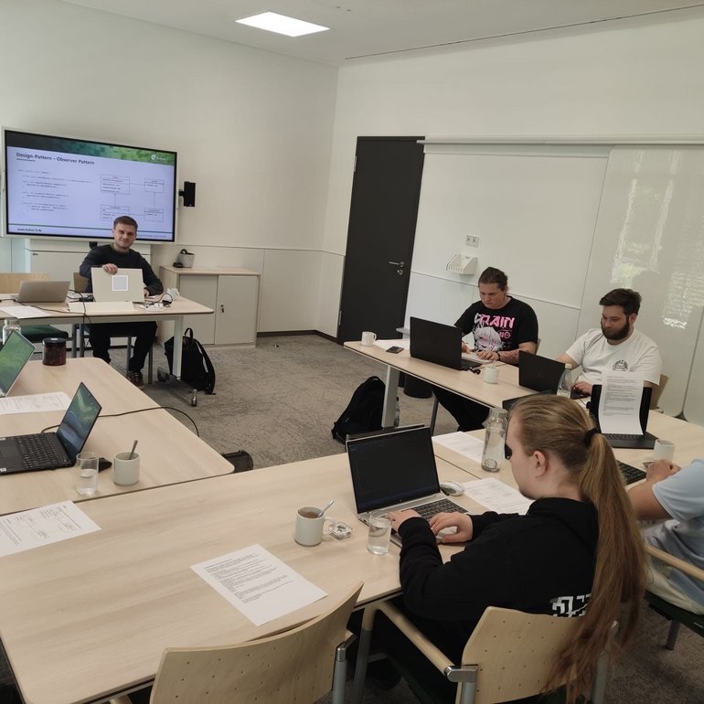 Das Bild zeigt eine Gruppe von Auszubildenden der kubus IT in einem modernen Seminarraum. Sie sitzen mit Laptops an Tischen und arbeiten konzentriert während eines Lehrgangs zur objektorientierten Programmierung. Im Hintergrund ist ein Dozent zu sehen, der Inhalte auf einem großen Bildschirm präsentiert. Die Atmosphäre wirkt fokussiert und praxisnah.