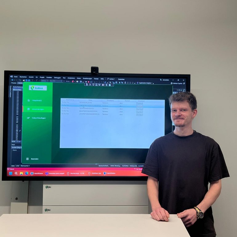 Der Auszubildende Philipp steht vor einem großen Bildschirm und präsentiert eine selbst entwickelte IT-Anwendung für Dokumentationsmanagement.