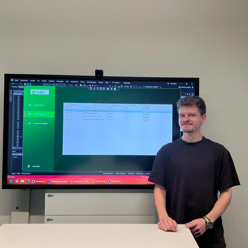 Der Auszubildende Philipp steht vor einem großen Bildschirm und präsentiert eine selbst entwickelte IT-Anwendung für Dokumentationsmanagement.