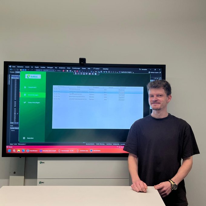 Der Auszubildende Philipp steht vor einem großen Bildschirm und präsentiert eine selbst entwickelte IT-Anwendung für Dokumentationsmanagement.