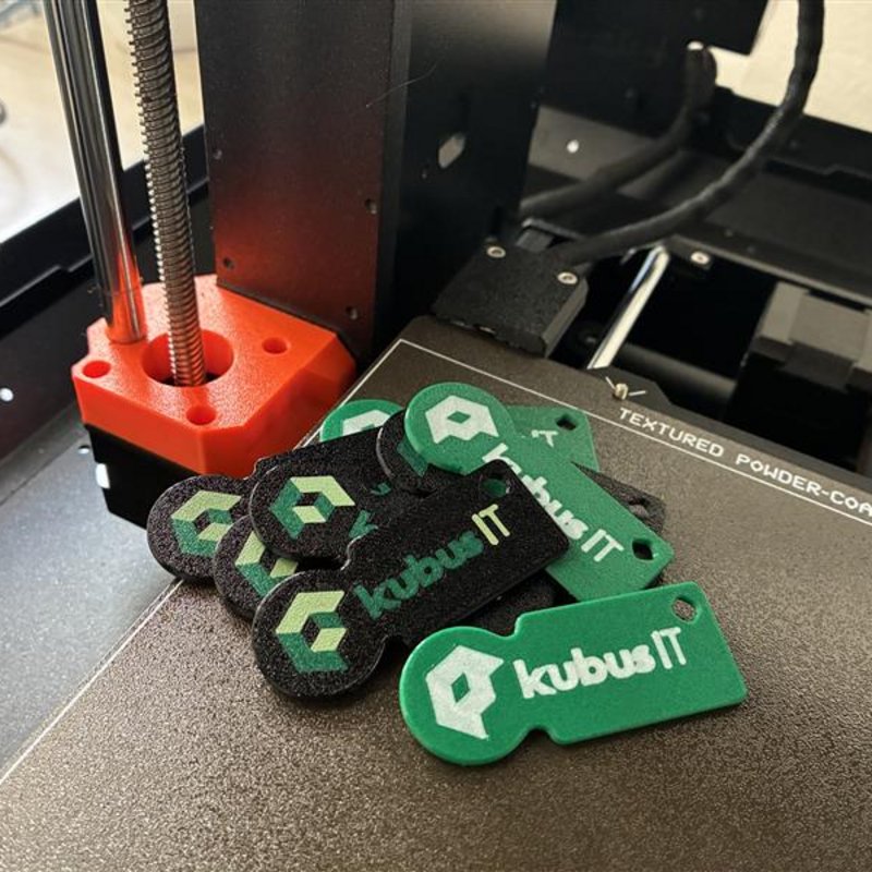 Mehrere grüne und schwarze Einkaufswagenchips mit kubus IT Logo liegen auf der Druckplatte eines 3D-Druckers.