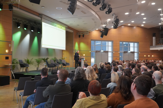 Eine weitwinklige Aufnahme aus dem Zuschauerraum im CongressCenter Erfurt. Im Fokus steht der Geschäftsführer der kubus IT, der auf einer Bühne an einem Rednerpult spricht. Er blickt in das Publikum, das in zahlreichen Stuhlreihen vor ihm sitzt und ihm de