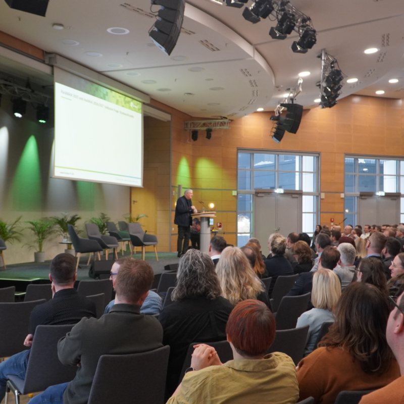 Eine weitwinklige Aufnahme aus dem Zuschauerraum im CongressCenter Erfurt. Im Fokus steht der Geschäftsführer der kubus IT, der auf einer Bühne an einem Rednerpult spricht. Er blickt in das Publikum, das in zahlreichen Stuhlreihen vor ihm sitzt und ihm den Rücken zuwendet. Auf der Bühne befindet sich links eine große Leinwand mit einer Präsentation sowie eine Sitzecke mit vier modernen grauen Sesseln und Grünpflanzen. Der Saal ist hell beleuchtet und verfügt über eine markante Holzvertäfelung an den Wänden.