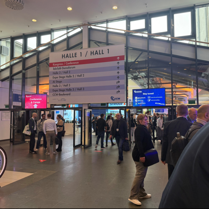 Eingangsbereich der CCW 2026 in Berlin mit Besucherandrang Im Eingangsbereich einer Messehalle stehen und gehen zahlreiche Besucherinnen und Besucher durch mehrere Glastüren. Über dem Eingang hängt ein großes Schild mit der Aufschrift „Halle 1 / Hall 1“ sowie Wegweisern zu Konferenz- und Ausstellungsbereichen. Digitale Bildschirme und Messestände sind im Hintergrund sichtbar, während sich Menschen in Business-Kleidung unterhalten und orientieren.