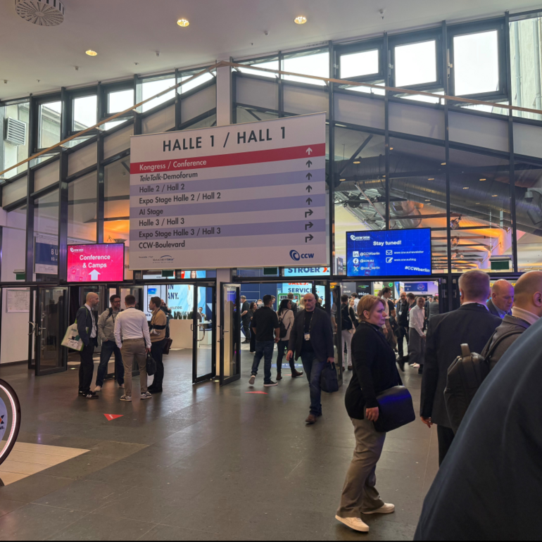 Im Eingangsbereich einer Messehalle stehen und gehen zahlreiche Besucherinnen und Besucher durch mehrere Glastüren. Über dem Eingang hängt ein großes Schild mit der Aufschrift „Halle 1 / Hall 1“ sowie Wegweisern zu Konferenz- und Ausstellungsbereichen. Digitale Bildschirme und Messestände sind im Hintergrund sichtbar, während sich Menschen in Business-Kleidung unterhalten und orientieren.