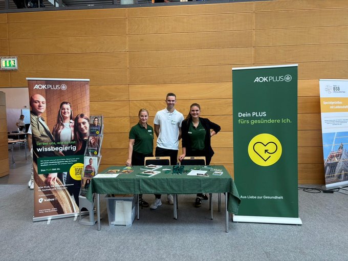 Drei Personen stehen hinter einem Messestand mit grünem Tischdecken, Flyern und Werbematerialien. Links und rechts sind große Roll-Ups von AOK PLUS mit den Botschaften „Sei bei uns, wie du bist: wissbegierig“ und „Dein PLUS fürs gesündere Ich“. Im Hintergrund ist eine moderne Messehalle mit Holzverkleidung zu sehen.