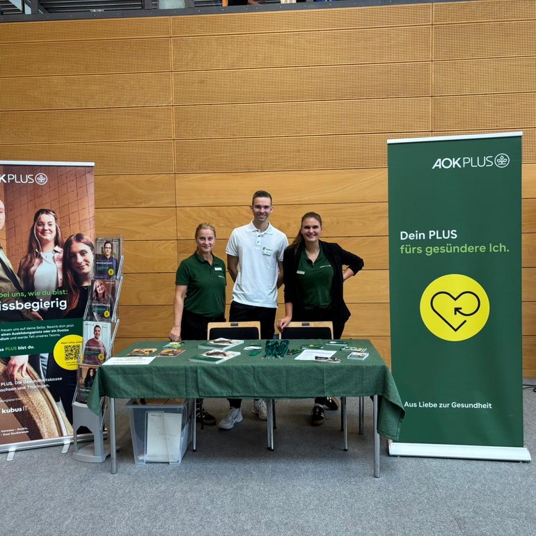 Drei Personen stehen hinter einem Messestand mit grünem Tischdecken, Flyern und Werbematerialien. Links und rechts sind große Roll-Ups von AOK PLUS mit den Botschaften „Sei bei uns, wie du bist: wissbegierig“ und „Dein PLUS fürs gesündere Ich“. Im Hintergrund ist eine moderne Messehalle mit Holzverkleidung zu sehen.