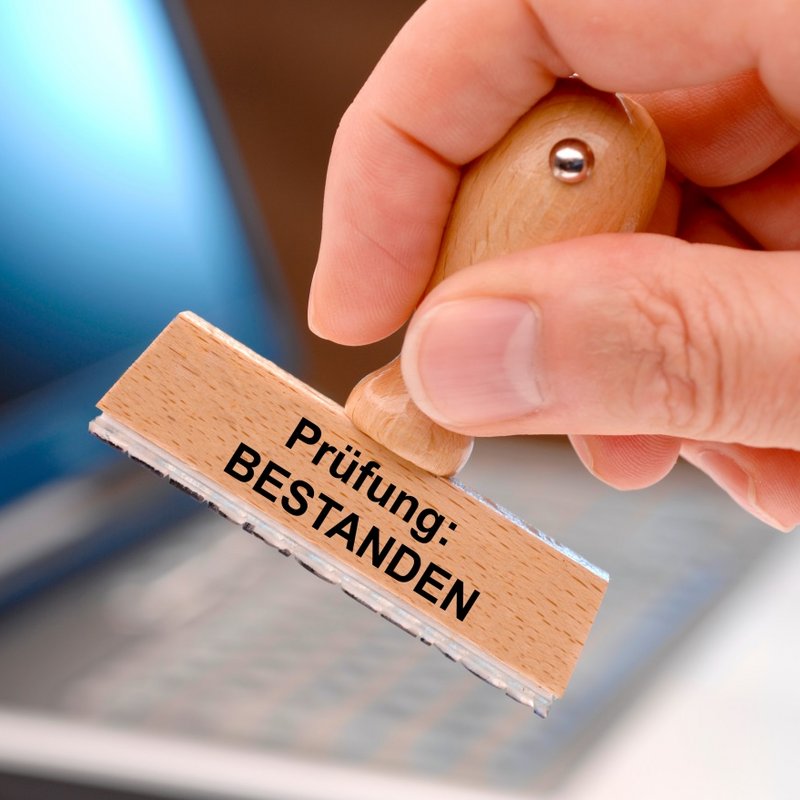 Stempel mit der Aufschrift „Prüfung: BESTANDEN“ wird von einer Hand über einem Laptop gehalten.