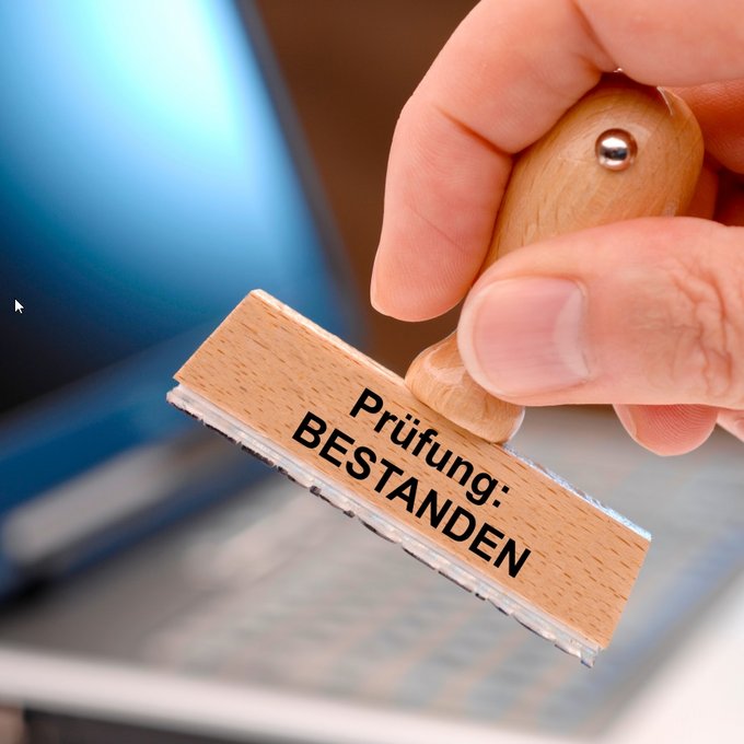 Eine menschliche Hand hält einen Holzstempel mit der Aufschrift „Prüfung: BESTANDEN“. Der Fokus liegt auf dem Stempel, der in den Vordergrund gehalten wird. Im Hintergrund ist unscharf ein aufgeklappter Laptop zu erkennen. Stempel mit der Aufschrift „Prüfung: BESTANDEN“ wird von einer Hand über einem Laptop gehalten.