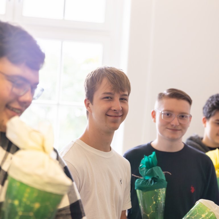 Auf dem Foto stehen drei junge Personen in einem hellen Raum mit großen Fenstern. Alle halten grüne Geschenktüten in Form von Zuckertüten in den Händen. Die linke Person trägt ein weißes T-Shirt und lächelt in Richtung Kamera. Die Person in der Mitte trägt ein dunkles Oberteil und blickt ebenfalls freundlich in die Kamera. Die rechte Person trägt einen dunkelgrauen Kapuzenpullover und betrachtet konzentriert die Zuckertüte in ihren Händen. Im Hintergrund sind Kartons zu sehen, die auf die Situation eines Büro- oder Schulungsraums hindeuten. Die Stimmung wirkt freundlich und entspannt.