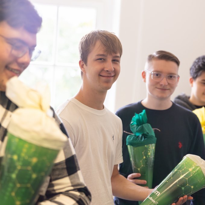 Auf dem Foto stehen drei junge Personen in einem hellen Raum mit großen Fenstern. Alle halten grüne Geschenktüten in Form von Zuckertüten in den Händen. Die linke Person trägt ein weißes T-Shirt und lächelt in Richtung Kamera. Die Person in der Mitte trägt ein dunkles Oberteil und blickt ebenfalls freundlich in die Kamera. Die rechte Person trägt einen dunkelgrauen Kapuzenpullover und betrachtet konzentriert die Zuckertüte in ihren Händen. Im Hintergrund sind Kartons zu sehen, die auf die Situation eines Büro- oder Schulungsraums hindeuten. Die Stimmung wirkt freundlich und entspannt.