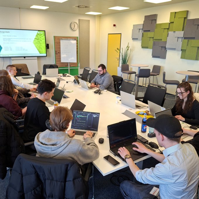 In einem hellen Seminarraum sitzen mehrere Auszubildende und Studierende an einem großen Tisch und arbeiten an ihren Laptops. An der Wand ist ein Bildschirm mit Präsentationsfolien zur Programmierung zu sehen. Die Teilnehmenden konzentrieren sich auf praktische Übungen und tauschen sich teilweise miteinander aus. Der Raum ist modern eingerichtet und für Gruppenarbeit ausgelegt.