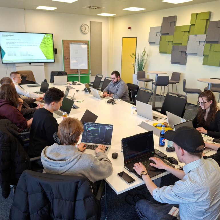 In einem hellen Seminarraum sitzen mehrere Auszubildende und Studierende an einem großen Tisch und arbeiten an ihren Laptops. An der Wand ist ein Bildschirm mit Präsentationsfolien zur Programmierung zu sehen. Die Teilnehmenden konzentrieren sich auf praktische Übungen und tauschen sich teilweise miteinander aus. Der Raum ist modern eingerichtet und für Gruppenarbeit ausgelegt.
