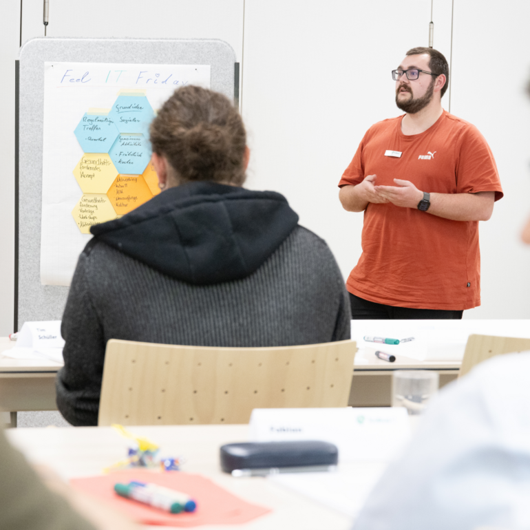 Ein junger Auszubildender in rotem Shirt präsentiert beim Talent Day 2025 der kubus IT im Bildungszentrum Hersbruck eine Gruppenarbeit, während Teilnehmende aufmerksam zuhören. Auf einem Flipchart stehen Themen wie Digitalisierung und Arbeitsumfeld.