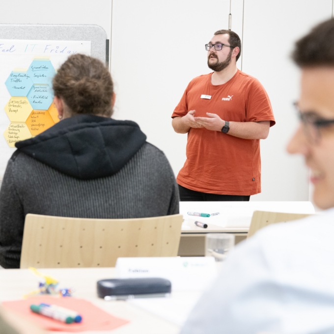 Talent Day 2025 der kubus IT: Austausch, Digitalisierung und Innovation im Fokus Ein junger Auszubildender in rotem Shirt präsentiert beim Talent Day 2025 der kubus IT im Bildungszentrum Hersbruck eine Gruppenarbeit, während Teilnehmende aufmerksam zuhören. Auf einem Flipchart stehen Themen wie Digitalisierung und Arbeitsumfeld.