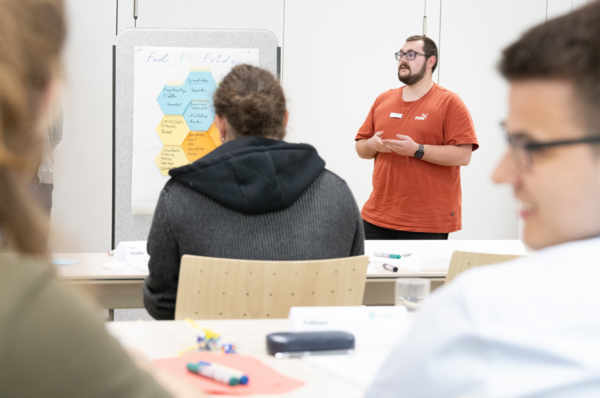 Ein junger Auszubildender in rotem Shirt präsentiert beim Talent Day 2025 der kubus IT im Bildungszentrum Hersbruck eine Gruppenarbeit, während Teilnehmende aufmerksam zuhören. Auf einem Flipchart stehen Themen wie Digitalisierung und Arbeitsumfeld.