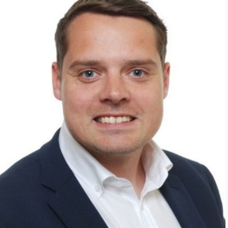 Das Bild zeigt Tom Blumenstein, den neuen Ausbildungsleiter der kubus IT. Er trägt ein Hemd, lächelt freundlich in die Kamera und steht vor einem neutralen Hintergrund.