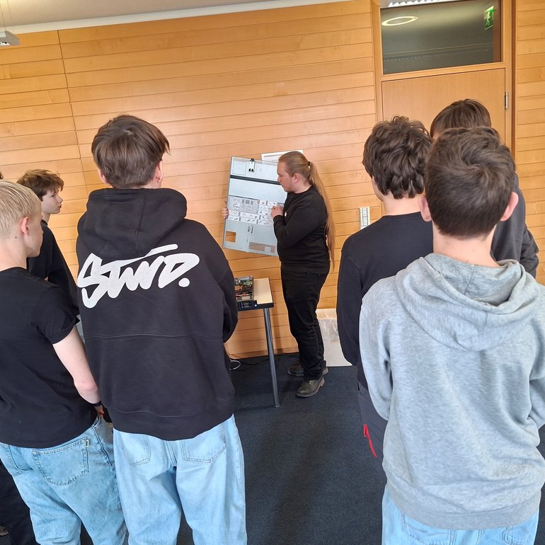 Eine Gruppe von Schülerinnen und Schülern steht in einem Raum und hört einer Person zu, die ein geöffnetes Computergehäuse erklärt und technische Komponenten zeigt