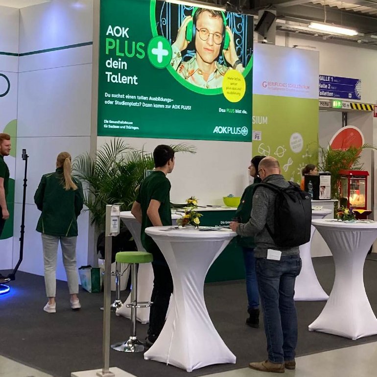 Einige Personen halten sich am Messestand auf, das Banner der AOK Plus ist im Hintergrund zu sehen