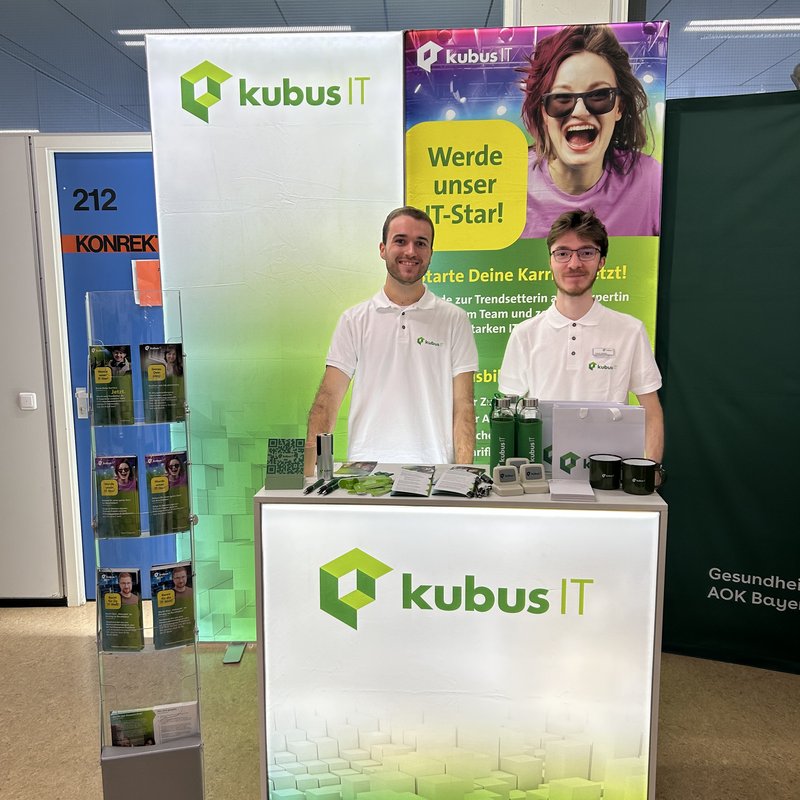Das Bild zeigt einen Messestand der kubus IT auf einer Ausbildungsmesse. Zwei Personen stehen hinter einem beleuchteten weißen Tresen mit dem kubus IT Logo in Grün. Beide tragen weiße Poloshirts mit dem Firmenlogo. Rechts auf dem Tresen liegen Werbematerialien wie Flyer, Kugelschreiber, Tassen und Trinkflaschen. Hinter ihnen befinden sich zwei große Banner: eines mit dem kubus IT Logo, das andere mit einem auffälligen Motiv einer jungen Person und dem Slogan „Werde unser IT-Star! Starte Deine Karriere jetzt!“. Links im Bild steht ein Prospektständer mit Flyern.