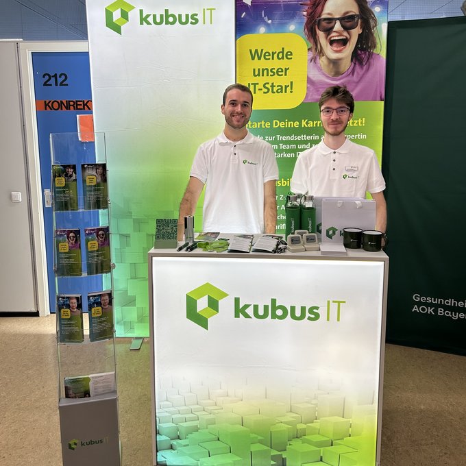 Das Bild zeigt einen Messestand der kubus IT auf einer Ausbildungsmesse. Zwei Personen stehen hinter einem beleuchteten weißen Tresen mit dem kubus IT Logo in Grün. Beide tragen weiße Poloshirts mit dem Firmenlogo. Rechts auf dem Tresen liegen Werbematerialien wie Flyer, Kugelschreiber, Tassen und Trinkflaschen. Hinter ihnen befinden sich zwei große Banner: eines mit dem kubus IT Logo, das andere mit einem auffälligen Motiv einer jungen Person und dem Slogan „Werde unser IT-Star! Starte Deine Karriere jetzt!“. Links im Bild steht ein Prospektständer mit Flyern.
