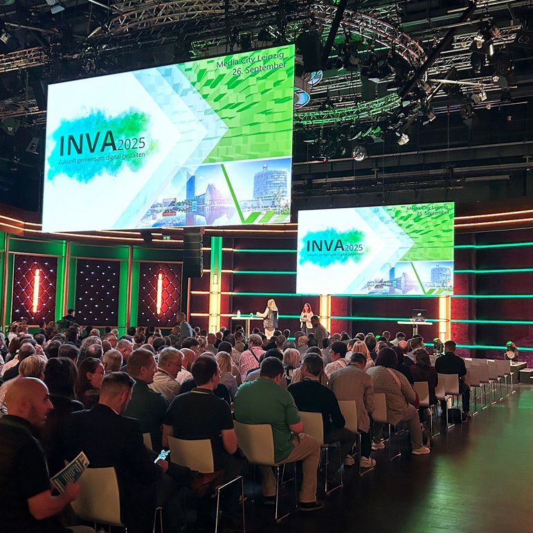 Großer Veranstaltungssaal mit Bühne und zwei großen Bildschirmen, auf denen „INVA 2025 – Zukunft gemeinsam digital gestalten“ angezeigt wird. Viele Menschen sitzen im Publikum und blicken nach vorne. Die Bühne ist in grün und orange beleuchtet, zwei Moderatorinnen stehen vor der Leinwand.