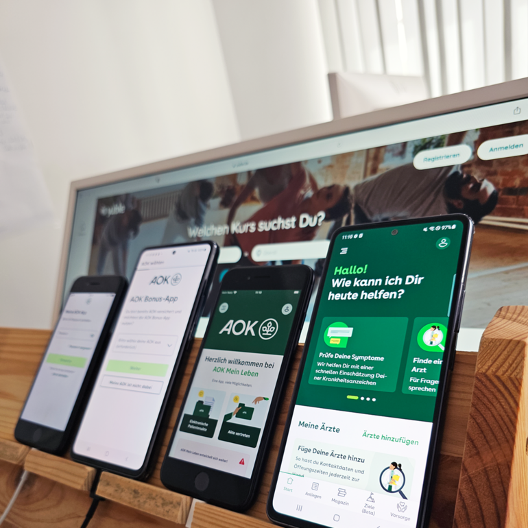 4 verschiedene Smartphones und im Hintergrund ein Monitor zeigen Apps der AOK