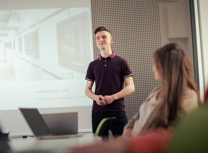 junger Mann hält eine Präsentation mithilfe eines Beamerbildes vor mehreren Personen im IT Gesundheitswesen | kubusIT - Arbeitsgemeinschaft von Körperschaften des öffentlichen Rechts eGbR