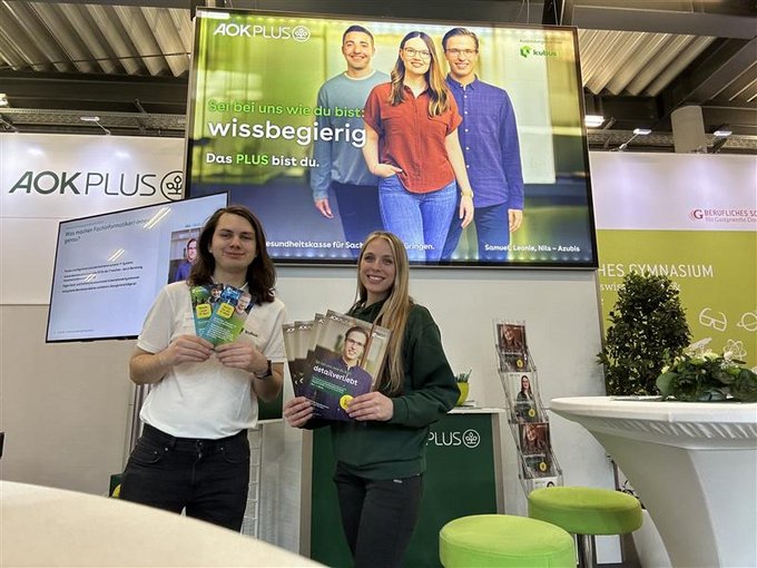 Ein Mann und eine Frau stehen lächelnd an einem Messestand der AOK PLUS und halten Informationsbroschüren in die Kamera. Im Hintergrund ist ein großer Monitor mit der Aufschrift „Sei bei uns wie du bist: wissbegierig. Das PLUS bist du.“ zu sehen, auf dem drei junge Menschen abgebildet sind. Der Stand ist modern gestaltet, mit grünen Hockern, Informationsmaterialien und dem Logo der AOK PLUS.