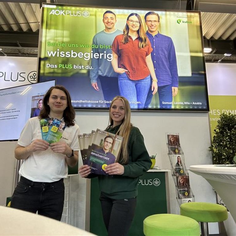 Ein Mann und eine Frau stehen lächelnd an einem Messestand der AOK PLUS und halten Informationsbroschüren in die Kamera. Im Hintergrund ist ein großer Monitor mit der Aufschrift „Sei bei uns wie du bist: wissbegierig. Das PLUS bist du.“ zu sehen, auf dem drei junge Menschen abgebildet sind. Der Stand ist modern gestaltet, mit grünen Hockern, Informationsmaterialien und dem Logo der AOK PLUS.