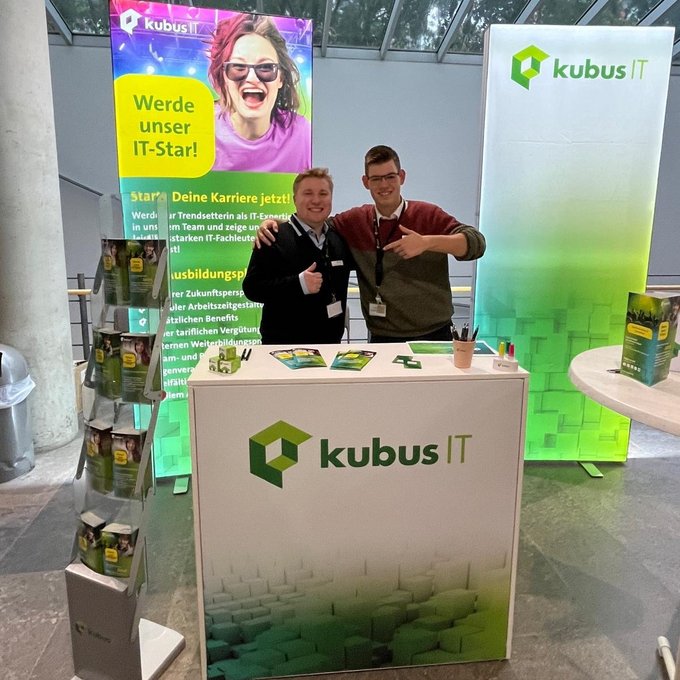 2 Mitarbeiter stehen am Messestand der kubus IT und lächeln in die Kamera
