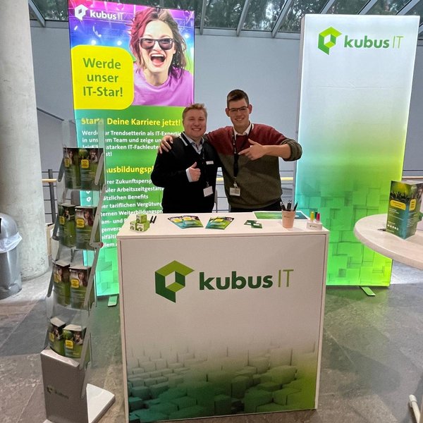 2 Mitarbeiter stehen am Messestand der kubus IT und lächeln in die Kamera
