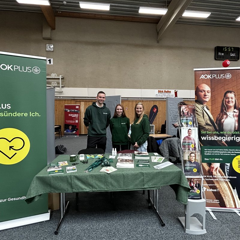 Zu sehen ist ein Messestand der AOK PLUS in einer Halle während einer Berufs- und Studienmesse. Mehrere Mitarbeitende stehen hinter einem Tisch mit Informationsmaterialien und Giveaways und sprechen mit Besucherinnen und Besuchern. Im Hintergrund sind Banner und Roll-ups mit Motiven aus dem Gesundheitswesen sowie weitere Messestände anderer Aussteller zu erkennen. Die Szene vermittelt eine offene, beratende Atmosphäre und zeigt den direkten Austausch mit interessierten Schülerinnen und Schülern.