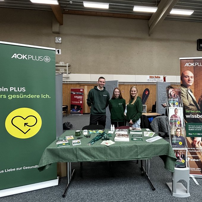Zu sehen ist ein Messestand der AOK PLUS in einer Halle während einer Berufs- und Studienmesse. Mehrere Mitarbeitende stehen hinter einem Tisch mit Informationsmaterialien und Giveaways und sprechen mit Besucherinnen und Besuchern. Im Hintergrund sind Banner und Roll-ups mit Motiven aus dem Gesundheitswesen sowie weitere Messestände anderer Aussteller zu erkennen. Die Szene vermittelt eine offene, beratende Atmosphäre und zeigt den direkten Austausch mit interessierten Schülerinnen und Schülern.
